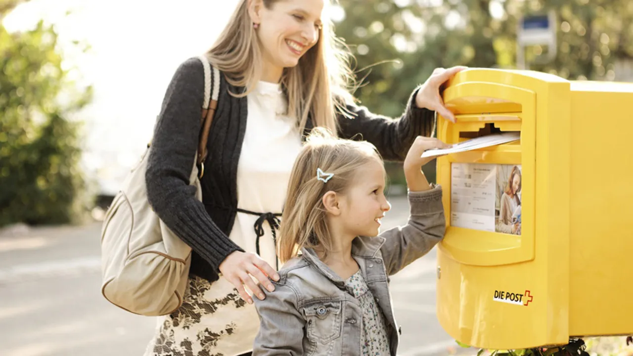 Mother and daughter posting a letter into a yellow Swiss Post mailbox outdoors