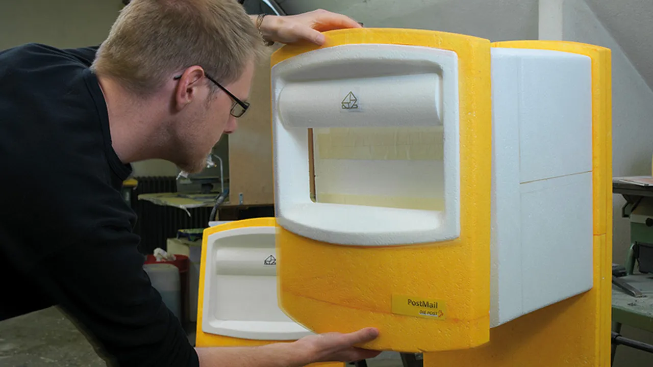 Technician examining yellow foam PostMail mailbox prototype inside workshop