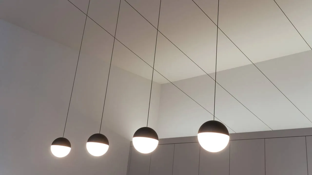Low-angle view of four modern spherical pendant lamps suspended from the ceiling, with dark upper halves and illuminated lower halves against a white ceiling grid.