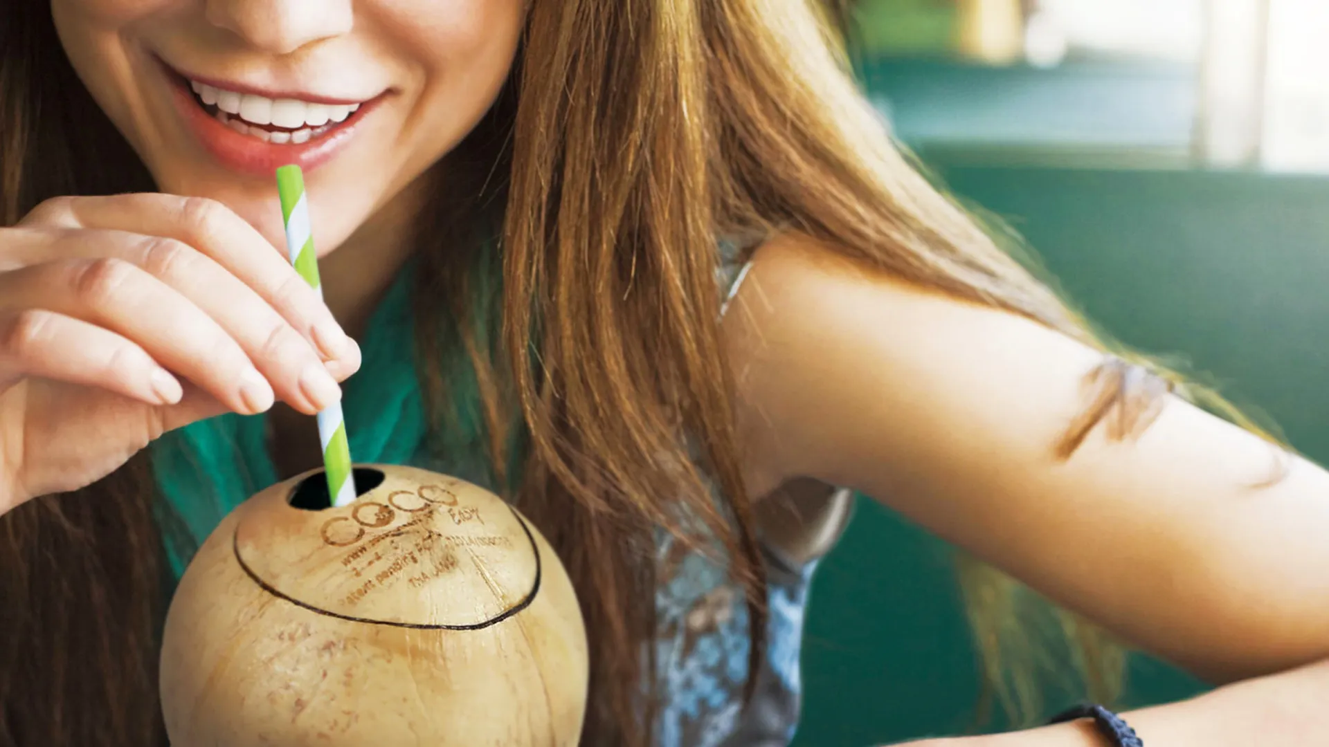 Close-up of a smiling woman drinking fresh coconut water through a green and white striped paper straw directly from the innovatively opened Coco Easy coconut.