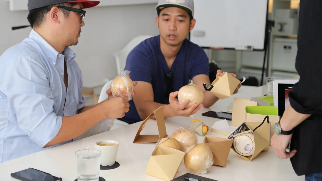 Two designers intently examining foil-wrapped coconuts and testing how they fit into the triangular kraft cardboard prototypes.