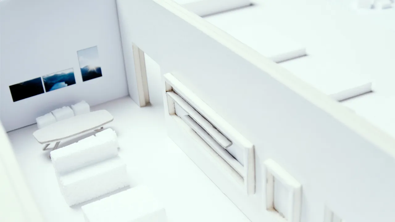 White architectural model of an interior space with miniature furniture, including a meeting table, armchairs, and tiny pictures on the walls.