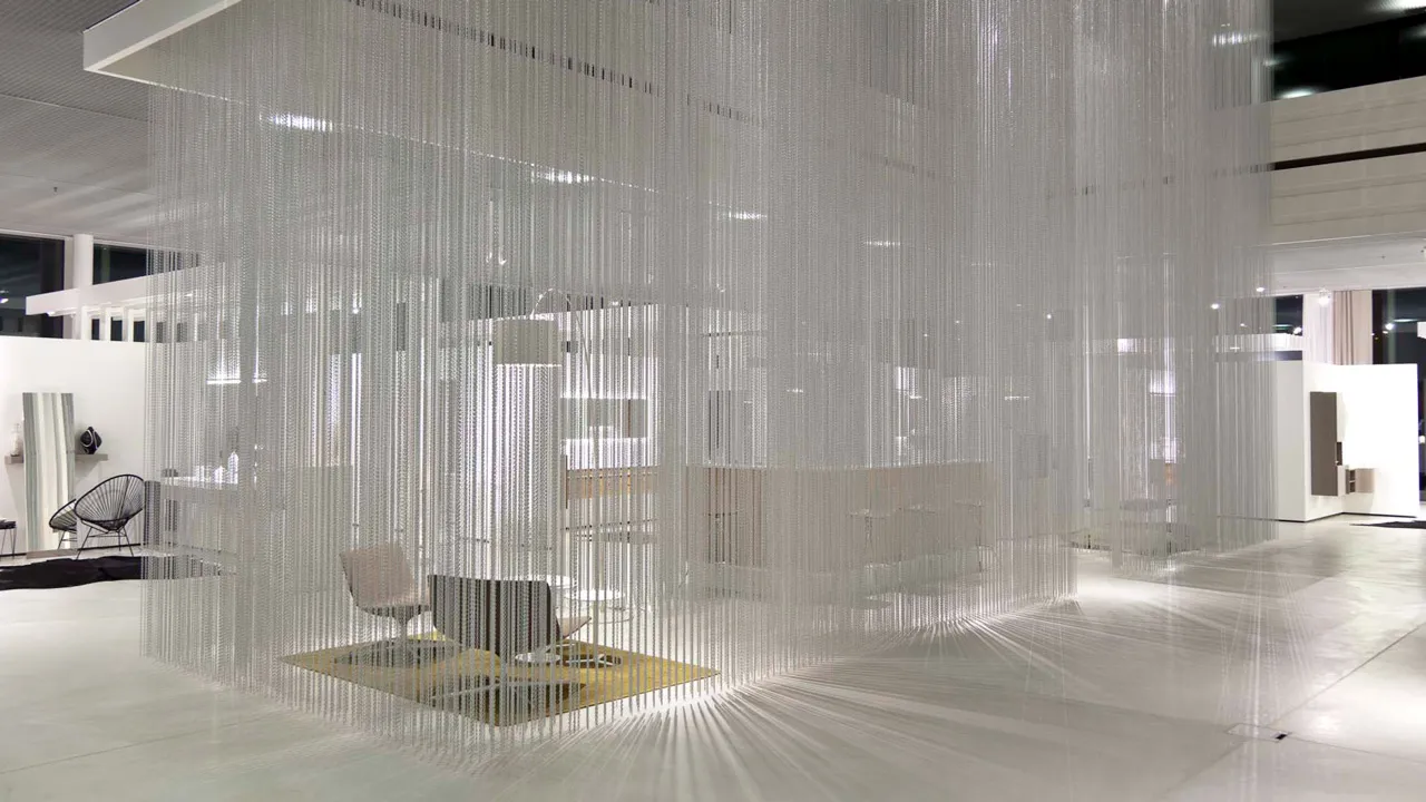 Interior shot of a bright showroom. A floor-to-ceiling installation made of thousands of fine metal chains forms a semi-transparent room around a seating area.