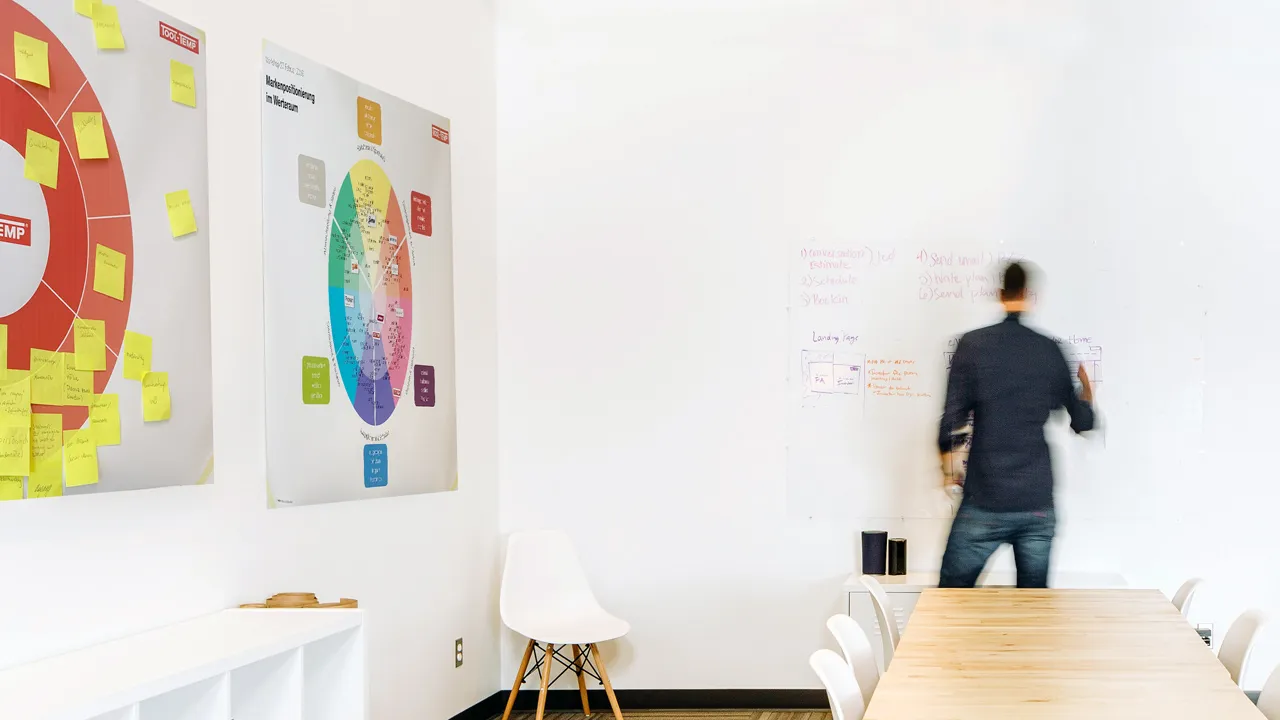 Helles Besprechungszimmer mit Holztisch, an dem ein Designer an einer Wand mit grossen Postern zur Markenpositionierung und einem Whiteboard arbeitet.